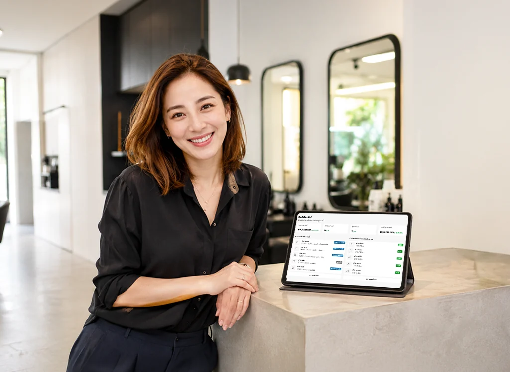 เจ้าของร้านเสริมสวยยืนอยู่ที่เคาน์เตอร์ ข้างแท็บเล็ตที่เปิดหน้าตารางคิวของ SalonX